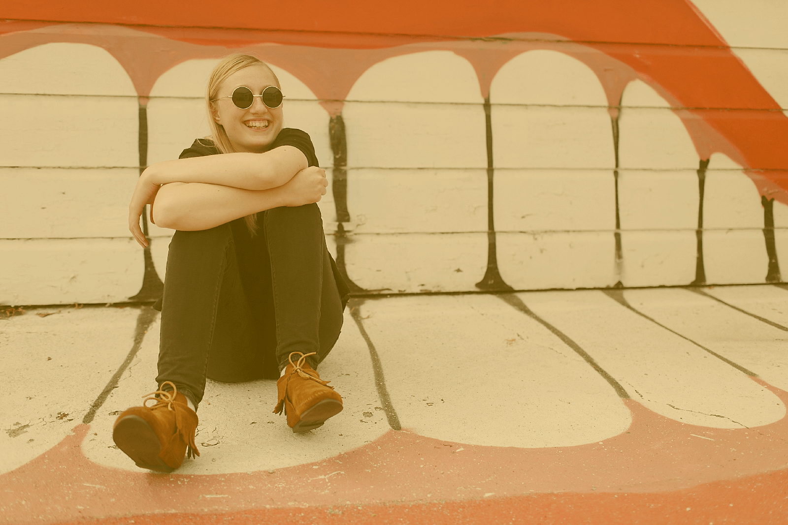 Eine junge Frau sitzt vor einem Garagentor auf dem Boden. Hinter und unter ihr befindet sich ein großes Grafiti von einem lachenden Mund.